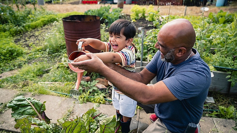 Un hombre ayuda a un niño sonriente a regar plantas en un jardín