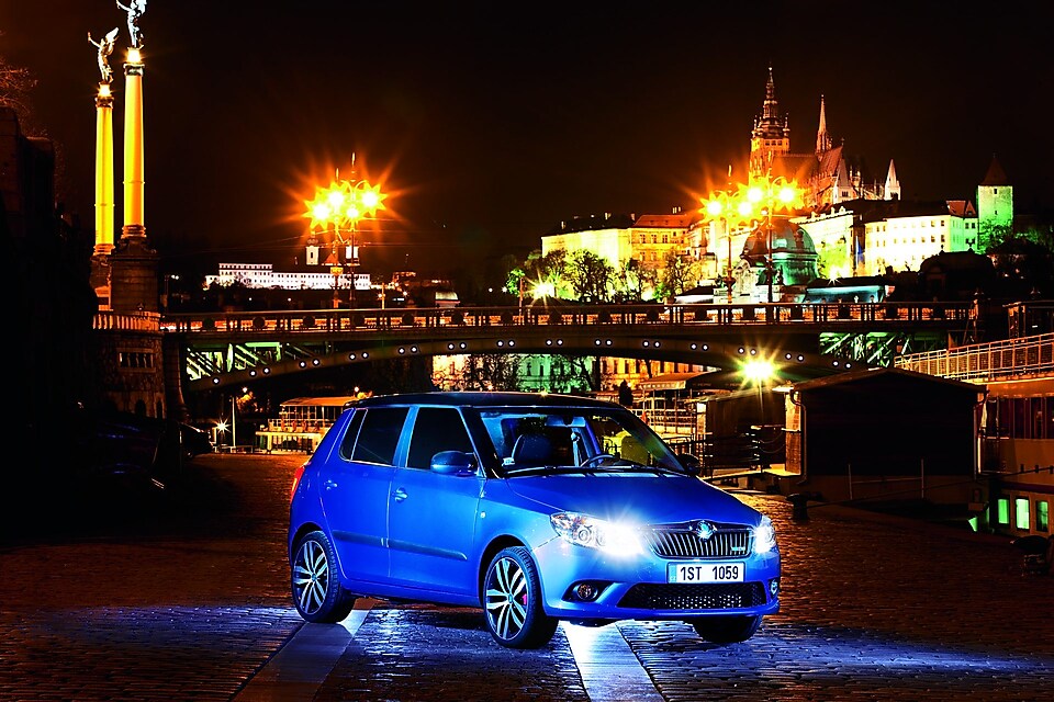 Coche azul aparcado en una calle de Praga con un puente y el castillo al fondo, de noche