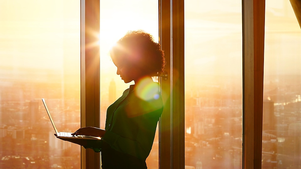 Mujer de pie junto a una ventana a contraluz de una puesta de sol utilizando un portátil