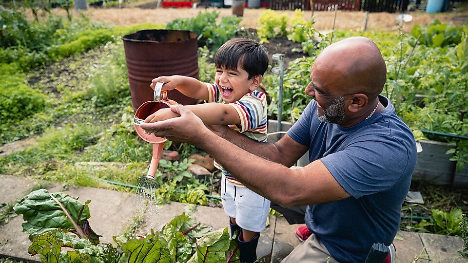 Un hombre ayuda a un niño sonriente a regar plantas en un jardín