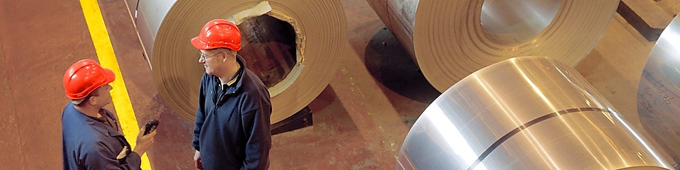 Two factory workers standing next to rolls of steel