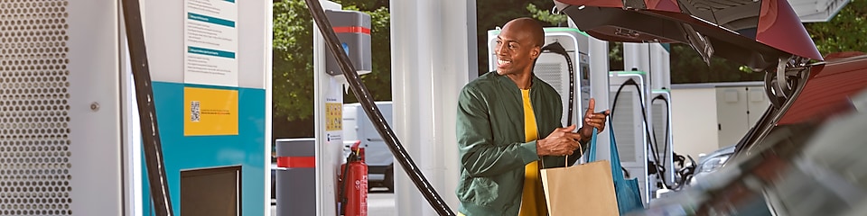 Señor sonriente en una estación de recarga de Shell junto a un vehículo con el maletero abierto en el que introduce unas bolsas.