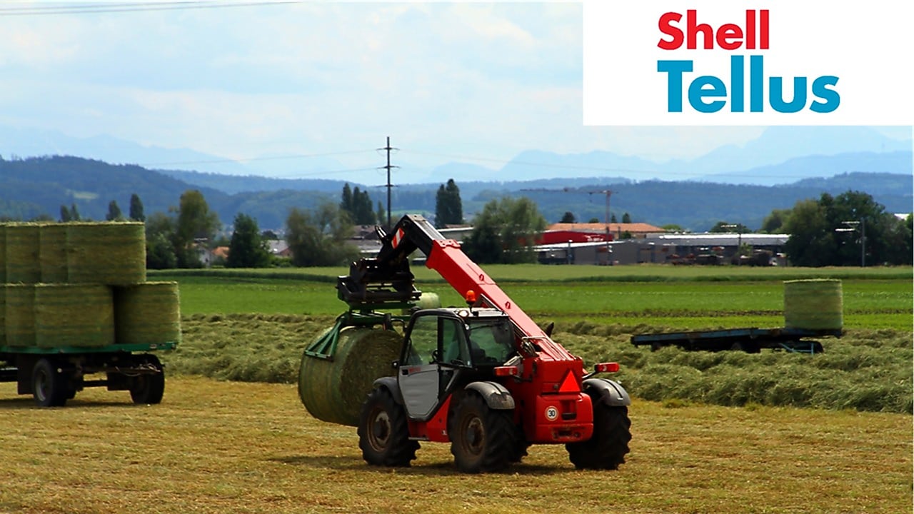 Un equipo agrícola con telescopio rojo levanta un gran fardo de heno redondo en un campo - Shell Tellus Logo