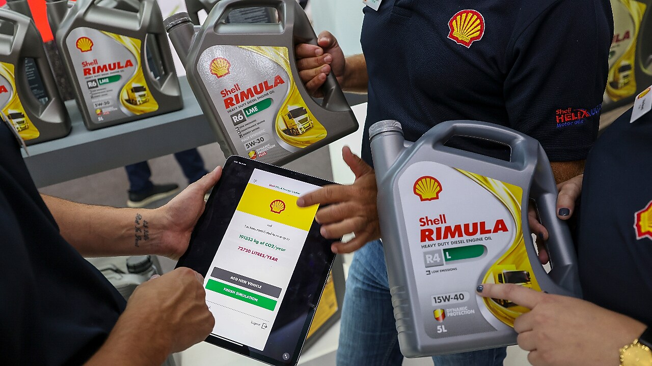 Tres personas sostienen botellas de Shell Rimula y una tablet que muestra el Shell CO₂ Energy Tracker. Al fondo, se observan estanterías con más botellas de Shell Rimula.