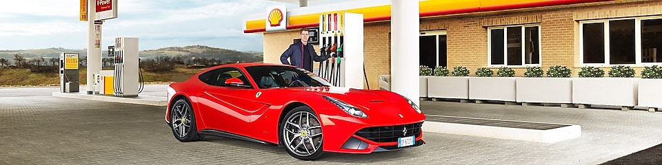 Ferrari rojo en una estación de servicio Shell con un hombre apoyado sobre un surtidor