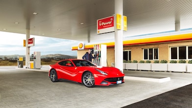 Ferrari rojo en una estación de servicio Shell con un hombre apoyado sobre un surtidor