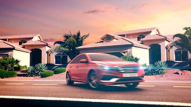 Coche rojo estacionado en el exterior de una casa con los faros encendidos al atardecer